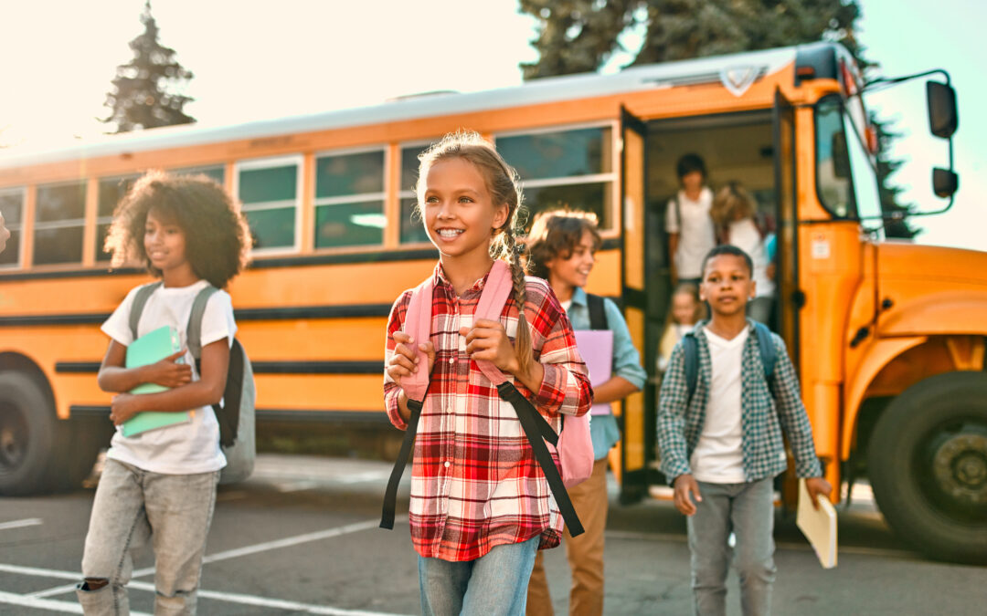 Students exiting a yellow school bus to walk across a parking lot into school. Students are wearing backpacks and carrying books or notebooks.