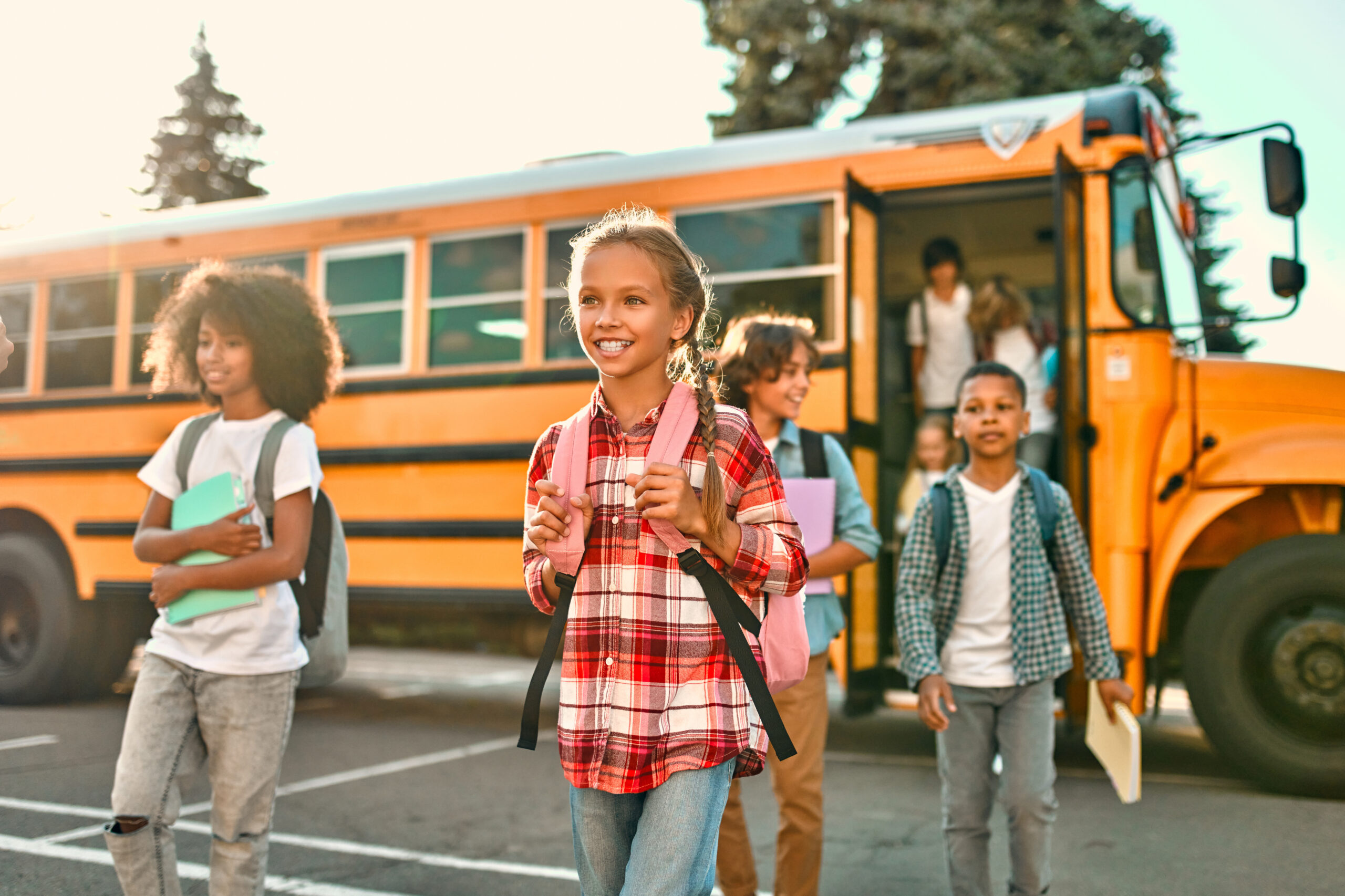 Students exiting a yellow school bus to walk across a parking lot into school. Students are wearing backpacks and carrying books or notebooks.